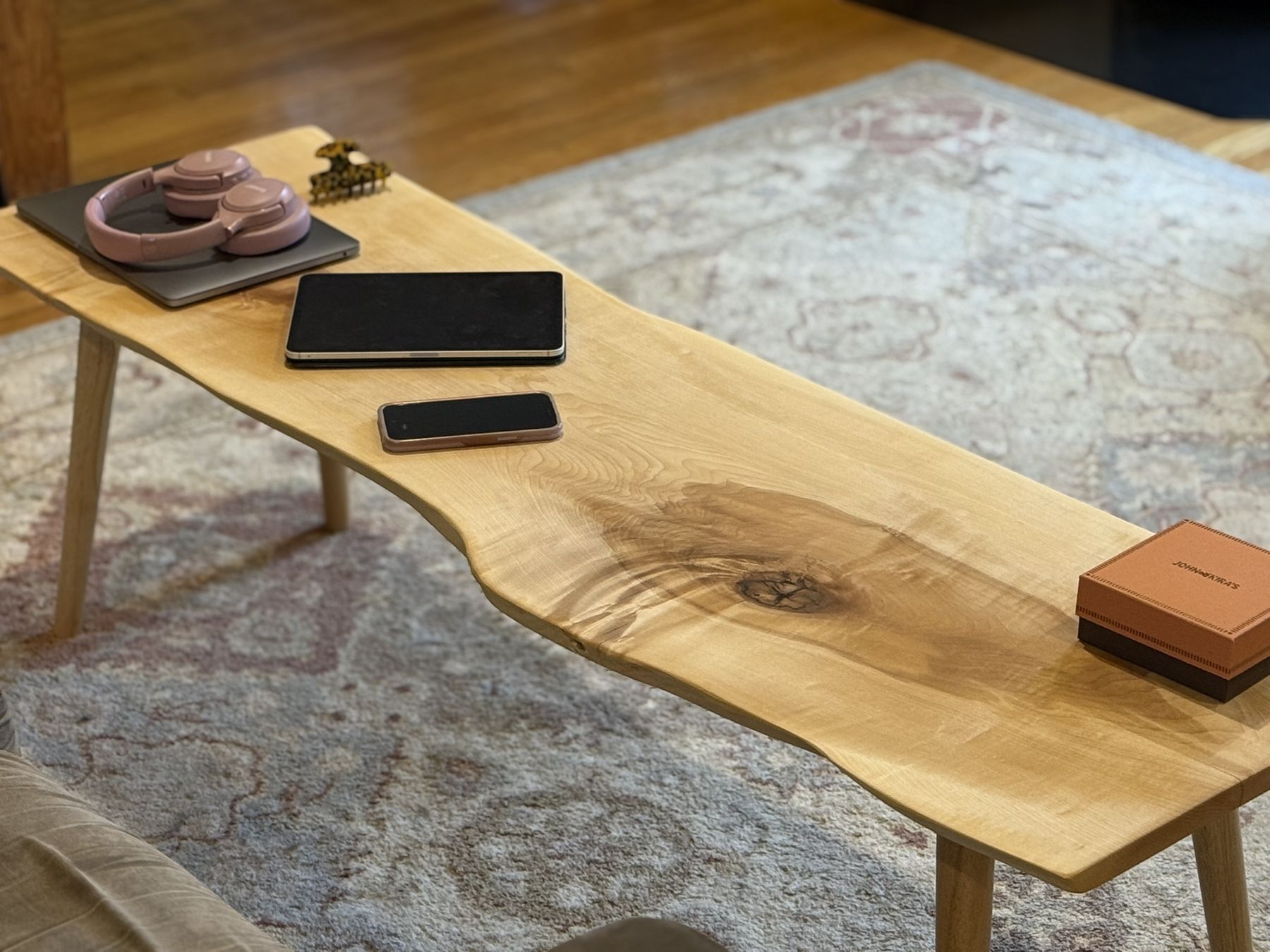 A long, pale live-edge coffee table on tapered round legs, with a laptop, tablet, phone, and headphones on top for scale.